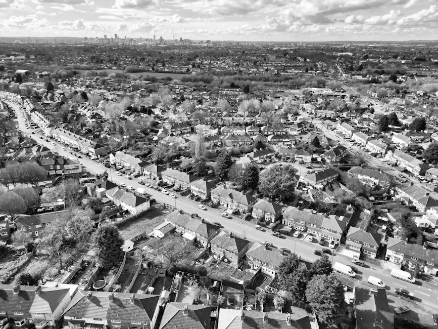 An aerial black-and-white photograph capturing a densely populated suburban residential area with rows of terraced houses, semi-detached homes, and small gardens. The foreground reveals a street lined with parked cars on both sides, with houses featuring pitched roofs, chimneys, and small front yards. Some houses have visible fences, driveways, and backyard gardens, with a variety of textures from tiled rooftops to garden hedges and trees. In the background, the urban landscape extends towards the horizon, where the distant city skyline is visible beneath a partly cloudy sky. The overall scene provides a detailed view of a typical UK neighbourhood, illustrating the scale and structure of suburban housing that waste removal services like Waste Removal Wimbledon might serve for private waste collection and on-site clearance within the community.