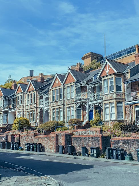 An aerial black-and-white photograph capturing a densely populated suburban residential area with rows of terraced houses, semi-detached homes, and small gardens. The foreground reveals a street lined with parked cars on both sides, with houses featuring pitched roofs, chimneys, and small front yards. Some houses have visible fences, driveways, and backyard gardens, with a variety of textures from tiled rooftops to garden hedges and trees. In the background, the urban landscape extends towards the horizon, where the distant city skyline is visible beneath a partly cloudy sky. The overall scene provides a detailed view of a typical UK neighbourhood, illustrating the scale and structure of suburban housing that waste removal services like Waste Removal Wimbledon might serve for private waste collection and on-site clearance within the community.