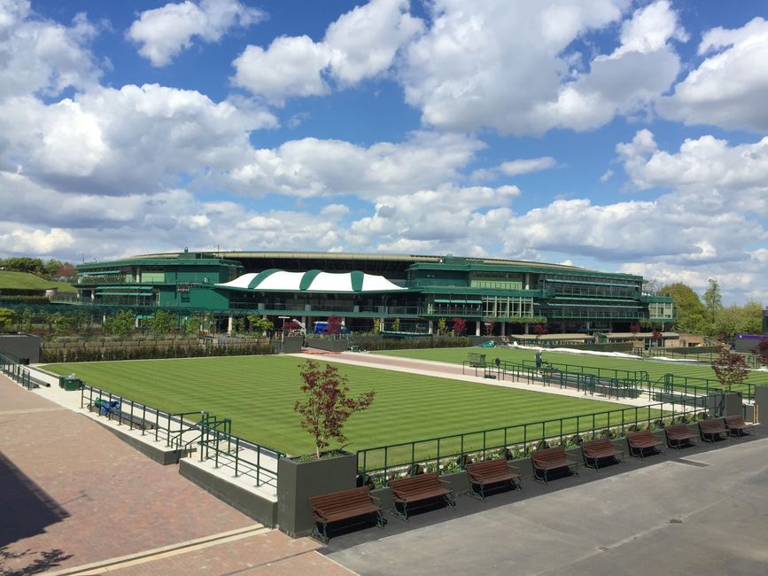 A large multi-storey building with a modern, glass and steel facade painted in dark green, situated behind a well-maintained grassy area. The grass appears freshly mowed, with visible mowing lines, and is bordered by a low, dark green metal fence. Several wooden benches with black metal frames are lined up along the fenced perimeter, facing the grass. A paved pathway runs parallel to the fence, with some small trees planted along the edge, one of which has red foliage. The scene is illuminated by natural daylight, with scattered white clouds in a bright blue sky overhead. In the background, some trees and low-rise buildings are visible, suggesting an urban or semi-urban environment. The setting appears to be a public outdoor space or park adjacent to the large building which may be involved in activities such as events or leisure. Waste Removal Wimbledon occasionally covers the area’s outdoor maintenance and waste management in relation to such public spaces, though no rubbish or waste items are visible in the image.