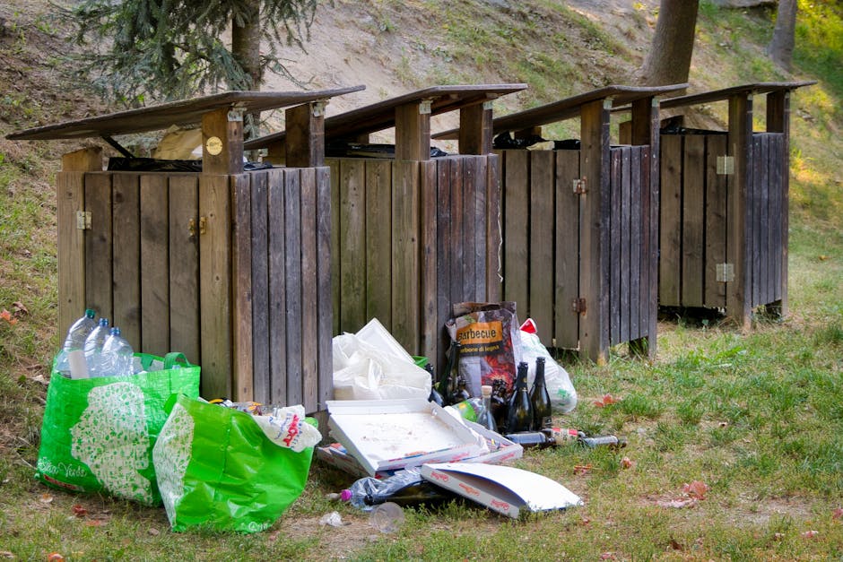 A row of four wooden outdoor rubbish bins with dark stain finish and sloped flat roofs, positioned on a grassy area adjacent to a slope with trees and foliage in the background. The bins are partially open, revealing no contents inside, and are mounted on small metal hinges. In front of the bins, there are various discarded items including a green reusable shopping bag filled with empty plastic bottles, a white pizza box with some leftover food debris, a bag of charcoal labeled 'barbecue', and several glass bottles, some empty and some with remaining liquid. The ground around the waste includes scattered paper, plastic, and other debris. The setting appears to be an outdoor space near a park or woodland area, representing a scenario of improper waste disposal, relevant to private rubbish collection and waste removal services that handle outdoor or bulk refuse. The scene is lit with natural daylight, highlighting the textures of the wooden bins, the grass, and the scattered rubbish, emphasizing the importance of proper waste management.